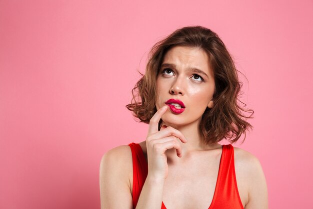 Portrait de gros plan de la pensée de la belle jeune femme aux lèvres rouges, toucher du doigt sa joue, regardant vers le haut