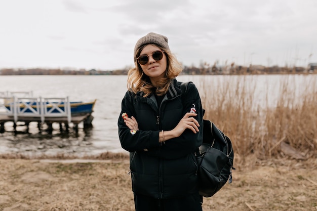 Portrait en gros plan en plein air d'une fille élégante et confiante en lunettes de soleil portant une casquette et une veste sombre met ses mains sur la poitrine contre le lac avec des bateaux