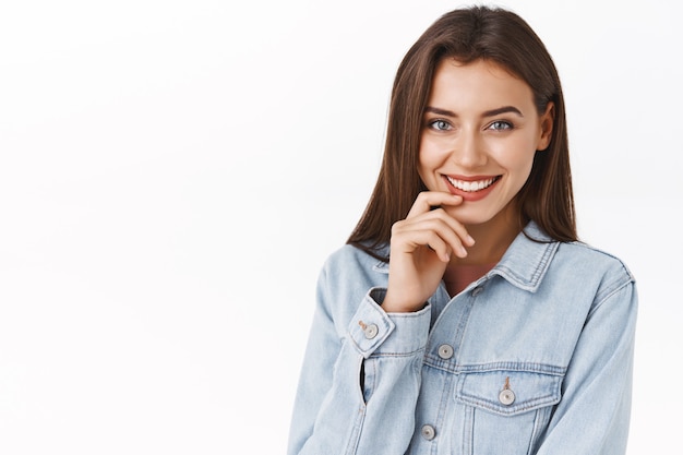 Portrait en gros plan séduisante femme sensuelle et féminine avec un joli sourire, une lèvre touchante et un rire coquette comme debout sur fond blanc en veste en jean, flirt express et séduction