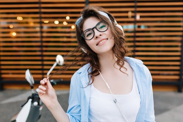 Portrait en gros plan d'une superbe fille joyeuse aux cheveux brun foncé jouant avec ses boucles et souriant timide