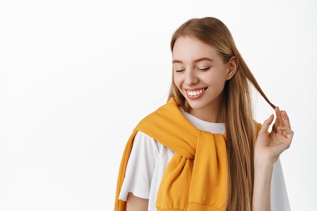 Portrait en gros plan d'une tendre fille blonde romantique, jouant avec une mèche de cheveux et évitant le contact visuel affectueux, riant et rougissant, souriant avec des dents blanches, debout contre le mur du studio