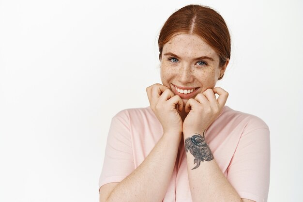 Portrait en gros plan, visage de jeune femme rousse avec une expression de visage mignon, taches de rousseur sur la peau du visage pâle, souriante coquette, ricanant affectueuse, debout sur blanc