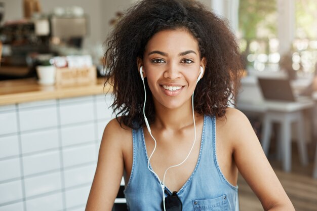 Portrait de l'heureuse belle femme africaine dans les écouteurs souriant assis dans le café.