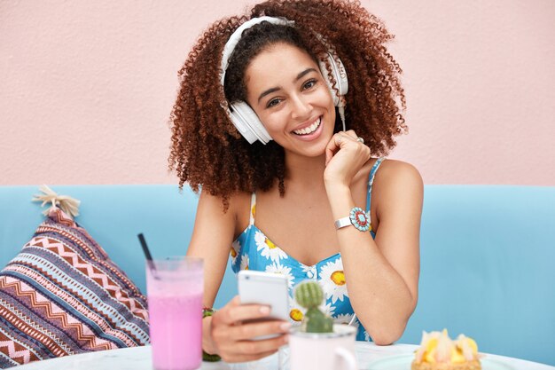 Portrait de l'heureuse jeune femme afro-américaine aux cheveux noirs croquants, écoute la radio, connecté à un téléphone intelligent moderne et un casque blanc