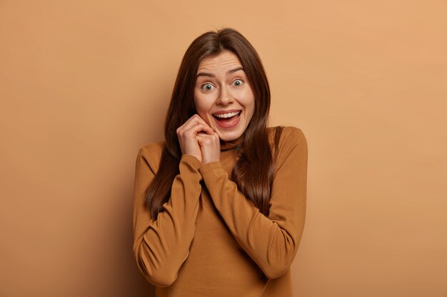 Portrait de l'heureuse jeune femme européenne a l'air ravie d'admiration et de joie, rit en recevant de bonnes nouvelles, porte un col roulé décontracté, pose contre un mur beige. Concept d'émotions