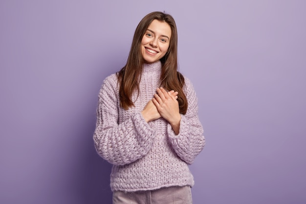 Portrait de l'heureuse jeune femme européenne garde les mains sur la poitrine, montre le geste du cœur, exprime sa gratitude, être reconnaissant, modèles contre le langage du corps mur violet. Monochrome. Les gens et la dévotion