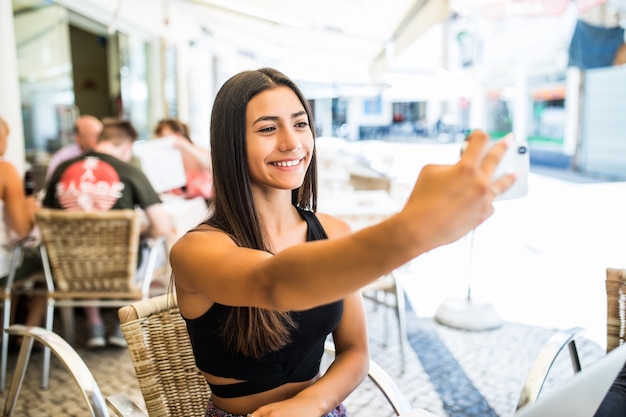 Portrait de l'heureuse jeune fille latine prenant selfie avec téléphone mobile alors qu'il était assis dans un café en plein air