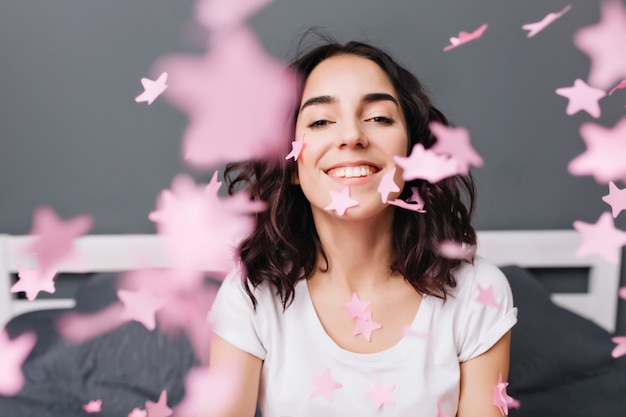 Portrait heureux excité jeune femme brune en T-shirt blanc s'amuser, souriant parmi les guirlandes roses tombant sur le lit dans un appartement moderne. Exprimer de vraies émotions positives, du bonheur