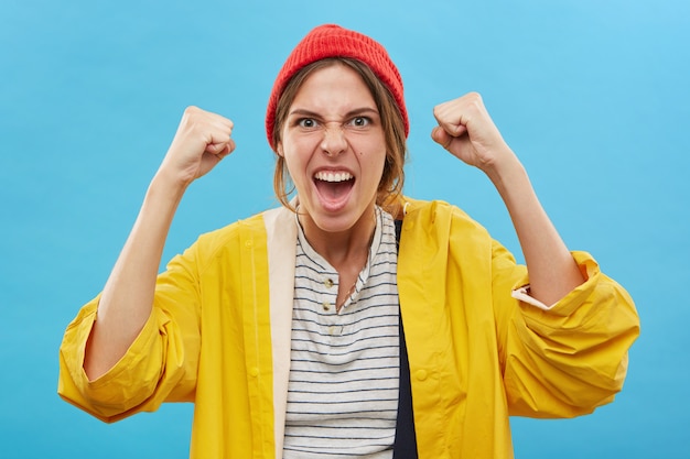 Photo gratuite portrait de l'heureux gagnant de la jeune femme caucasienne réussie portant un chapeau rouge et un imperméable jaune se réjouissant de la victoire, du succès ou de bonnes nouvelles positives avec les poings fermés, acclamant, criant de joie