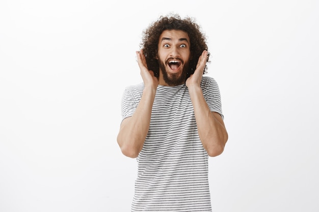 Portrait d'heureux mannequin homme beau débordé avec barbe et cheveux bouclés, hurlant d'émotions surprises et positives, tenant les paumes près du visage