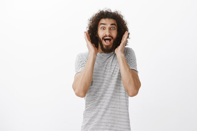 Portrait d'heureux mannequin homme beau débordé avec barbe et cheveux bouclés, hurlant d'émotions surprises et positives, tenant les paumes près du visage