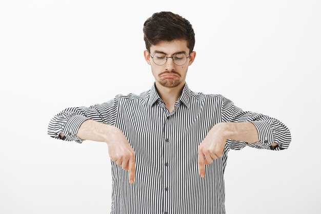 Portrait de heureux mec barbu attrayant calme en chemise rayée, pointant et regardant vers le bas avec une expression triste sombre, être déçu et contrarié par un résultat négatif sur un mur gris