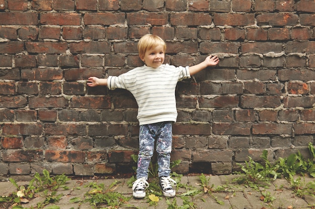 Photo gratuite portrait d'heureux petit garçon insouciant. petit enfant debout près du mur de briques, étirant les bras à l'extérieur. il porte un t-shirt rayé, un jean décontracté avec des taches colorées, des chaussures en caoutchouc grunge. les pissenlits poussent autour.