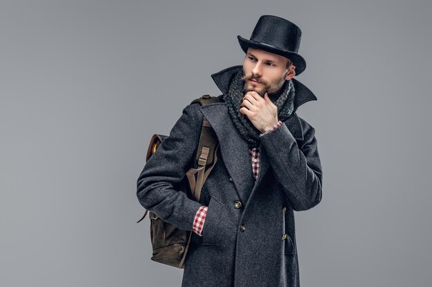 Portrait d'un hipster barbu vêtu d'une veste grise et d'un chapeau cylindrique tenant un sac à dos isolé sur fond gris.