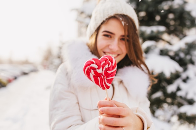 Portrait hiver jeune femme tenant une sucette coeur rose, se détendre sur la rue pleine de neige en matinée ensoleillée. Bonnet tricoté blanc, souriant.