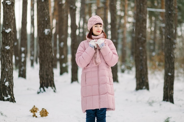 Portrait d'hiver en plein air d'une jolie jeune femme portant un manteau rose et un chapeau et une écharpe tricotés élégants, debout dans une belle forêt enneigée devant des pins et profitant d'une promenade par temps glacial.