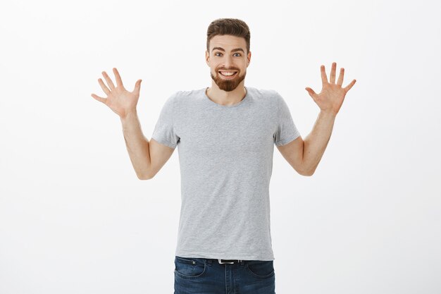 Portrait d'homme adulte beau heureux excité et ravi avec barbe, moustache et yeux bleus levant les paumes montrant dix doigts et souriant largement étonné de dire des nouvelles impressionnantes contre le mur gris