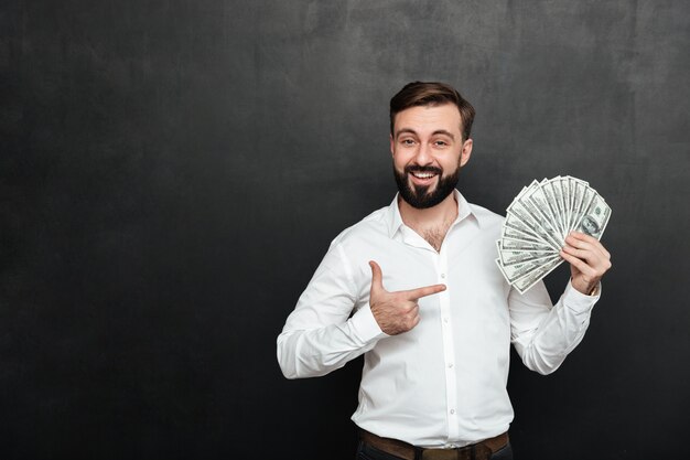 Portrait d'homme adulte en chemise blanche se présentant à la caméra avec ventilateur de billets de 100 dollars à la main, étant riche et heureux sur gris foncé