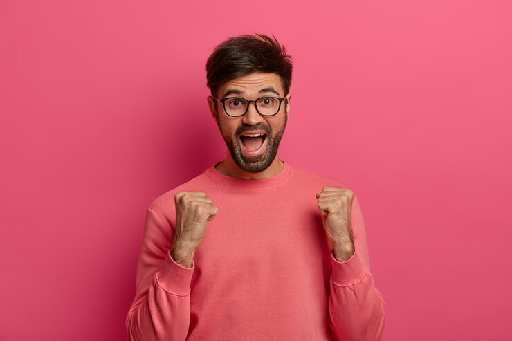 Photo gratuit portrait d'un homme adulte mal rasé joyeux serre les poings avec triomphe, se réjouit du bon moment de la vie, s'exclame positivement, porte des lunettes transparentes et un pull rose, célèbre la victoire ou le succès