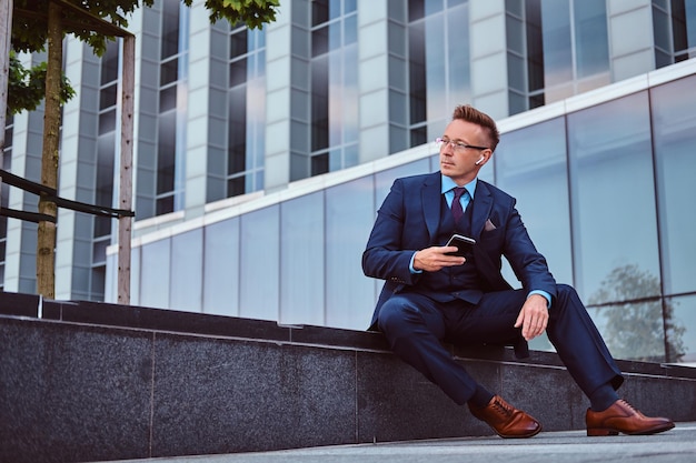 Photo gratuite portrait d'un homme d'affaires élégant et confiant vêtu d'un costume élégant tenant un smartphone et regardant ailleurs tout en étant assis à l'extérieur sur fond de gratte-ciel.