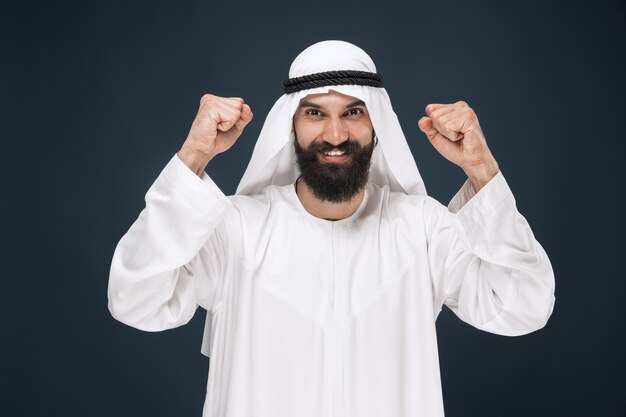 Portrait d'homme d'affaires saoudien arabe sur fond de studio bleu foncé. Jeune mannequin debout, souriant et célébrant. Concept d'entreprise, finance, expression faciale, émotions humaines.