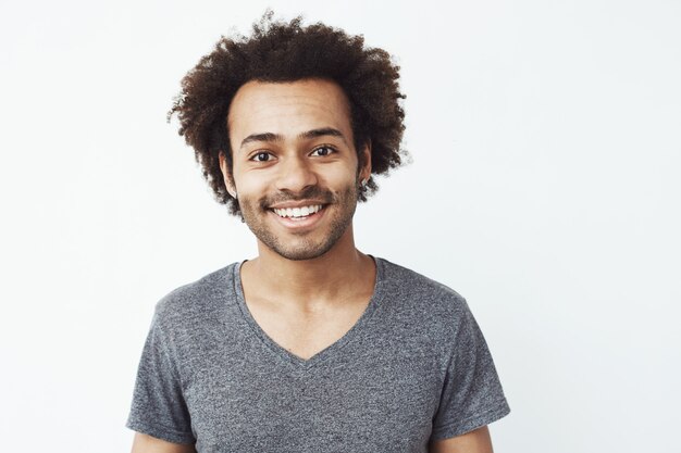 Portrait d'un homme africain heureux et charmant souriant, un petit ami en attente d'une date, ou un rêve de chasseur de têtes debout sur un mur blanc.