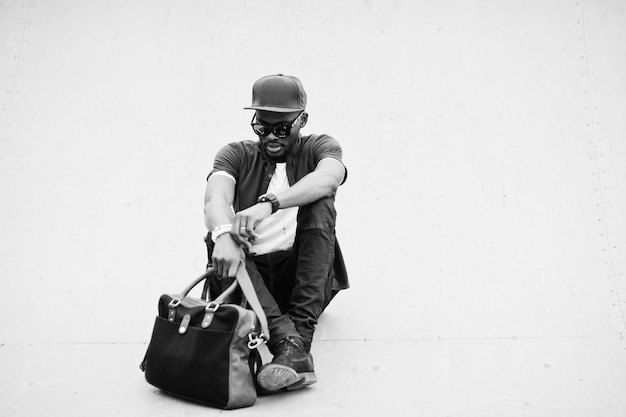 Portrait d'un homme afro-américain élégant assis portant des lunettes de soleil et une casquette avec sac à main en plein air Street fashion black man