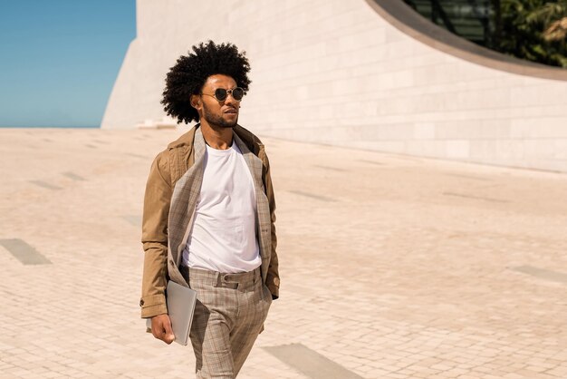 Portrait d'homme afro-américain à la mode à l'extérieur. Homme en costume avec barbe tenant un ordinateur portable, marchant. Portrait, concept de vie urbaine