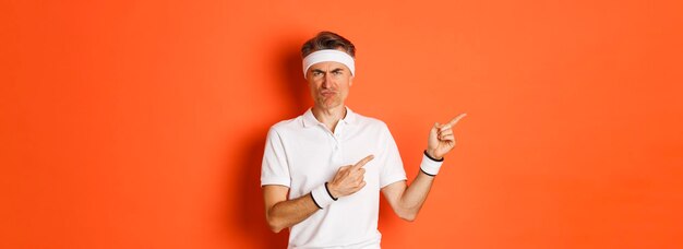 Portrait d'un homme d'âge moyen déçu en vêtements de sport se plaignant et pointant les doigts vers la gauche sur le logo montrant quelque chose de mal debout sur fond orange