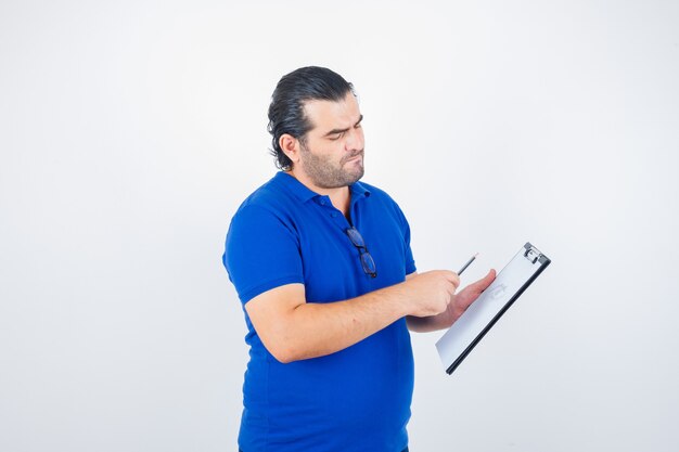 Portrait d'un homme d'âge moyen regardant à travers le presse-papiers tout en tenant un crayon en polo t-shirt et à la pensif