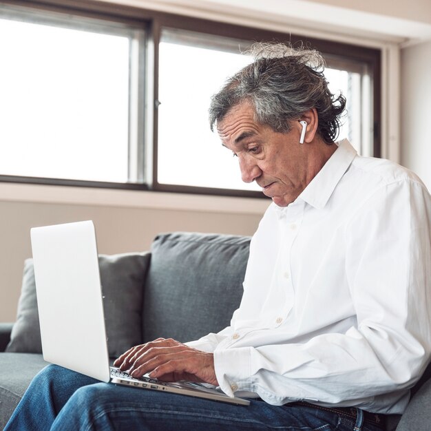 Portrait, homme aîné, séance, sofa, porter, écouteur bluetooth, utilisation, ordinateur portable