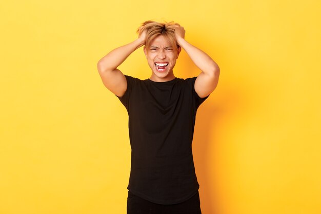 Portrait d'un homme asiatique en colère énervé jetant la coupe de cheveux et hurlant furieux, debout sur un mur jaune