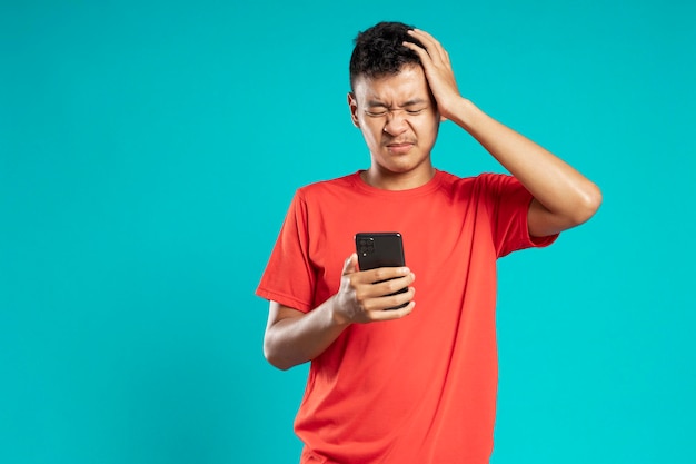 Portrait d'un homme asiatique étourdi parlant sur smartphone et tenant la tête isolée sur fond bleu