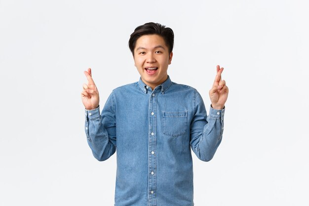 Portrait d'un homme asiatique optimiste et plein d'espoir avec des bretelles, souriant optimiste, ayant foi dans les rêves devenus réalité. Guy faisant un vœu avec les doigts croisés, anticipant un miracle, se sentant chanceux, fond blanc.