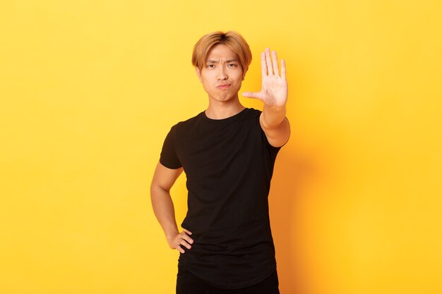 Portrait d'un homme asiatique sérieux déçu, souriant mécontent et étendre la main, montrant le geste d'arrêt, mur jaune