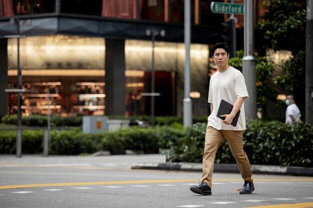 Portrait d'un homme asiatique traversant la rue de la ville tout en tenant un ordinateur portable