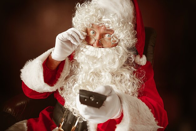 Portrait d'homme au père Noël avec une barbe blanche luxueuse, un chapeau du père Noël et un costume rouge au fond de studio rouge