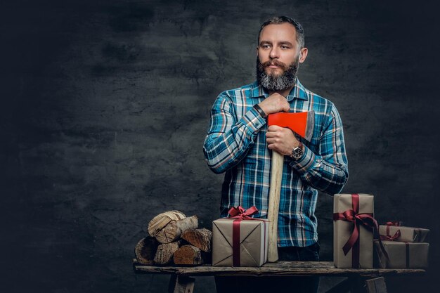 Portrait d'un homme barbu d'âge moyen tenant une hache et debout près d'une table avec des coffrets cadeaux de Noël et des bois de chauffage sur fond gris.