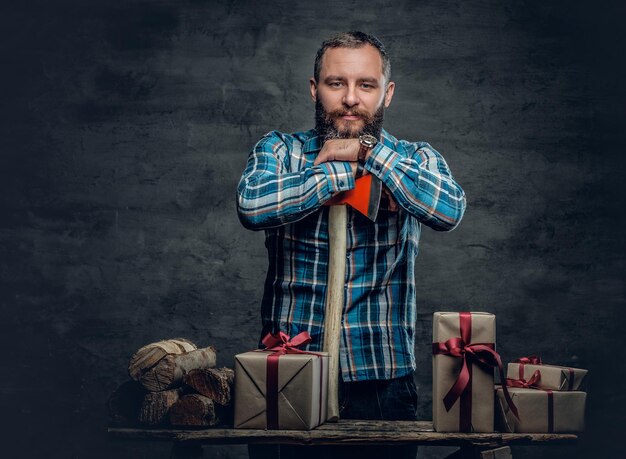 Portrait d'un homme barbu d'âge moyen tenant une hache et debout près d'une table avec des coffrets cadeaux de Noël et des bois de chauffage sur fond gris.