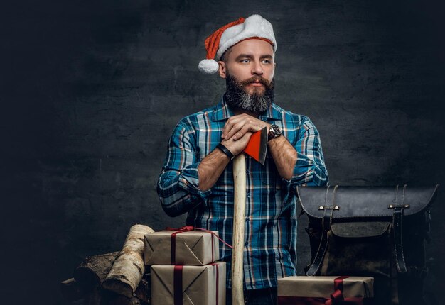 Portrait d'un homme barbu d'âge moyen vêtu d'une chemise en flanelle à carreaux et d'un chapeau de Père Noël tenant une hache et debout près d'une table avec des coffrets cadeaux de Noël et des bois de chauffage sur fond gris.
