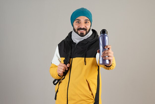 Portrait d'homme barbu au chapeau chaud debout et tenant une bouteille d'eau contre un mur gris.