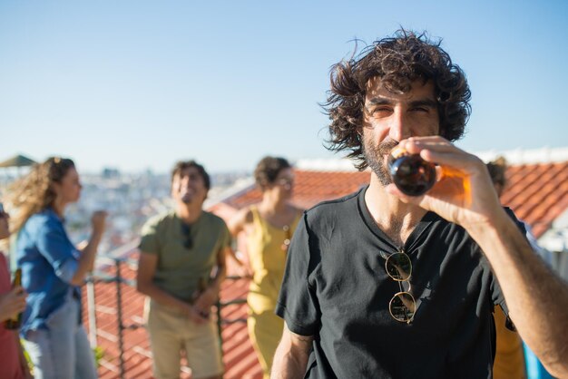 Portrait d'homme barbu aux cheveux bouclés à la fête. Jeune homme de race blanche en T-shirt noir buvant de la bière. Amis de différentes nationalités en arrière-plan. Fête, concept d'amitié