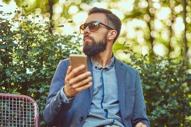 Portrait d'un homme barbu avec une coupe de cheveux élégante dans des vêtements décontractés, écrit un message au téléphone tout en étant assis sur un banc dans un parc de la ville.