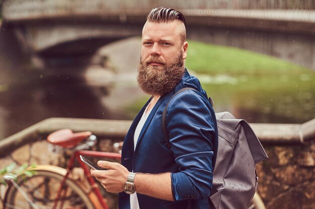 Portrait d'un homme barbu avec une coupe de cheveux vêtu de vêtements décontractés avec un sac à dos, debout dans un parc de la ville, à l'aide d'un smartphone.