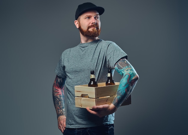 Portrait d'homme barbu dans une casquette avec des tatouages sur ses bras tient une boîte en bois avec des bouteilles de bière.