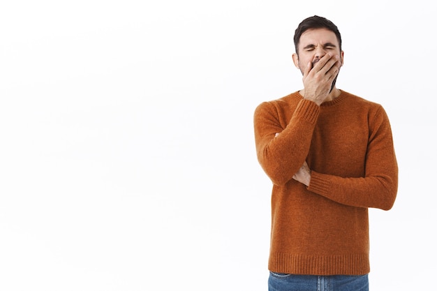 Portrait D'un Homme Barbu Fatigué Se Sentant épuisé Après Avoir Travaillé Tard Dans La Nuit, Bâillant Et Couvrant La Bouche Avec La Paume, Debout Ennuyé Ou Endormi, Attendant Une Tasse De Café Le Matin Au Bureau, Mur Blanc