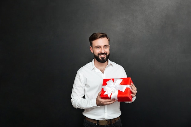 Portrait d'un homme barbu heureux tenant une boîte-cadeau rouge et regardant la caméra sur un mur gris foncé