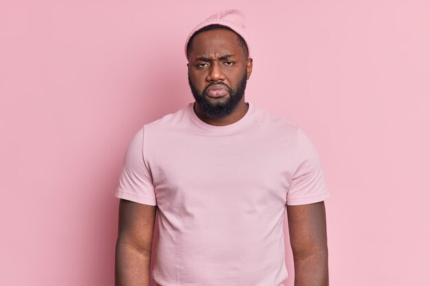 Portrait d'homme barbu mécontent frustré regarde malheureusement la caméra d'être insatisfait de quelque chose porte un chapeau et un t-shirt décontracté isolé sur un mur rose