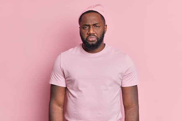 Portrait d'homme barbu mécontent frustré regarde malheureusement la caméra d'être insatisfait de quelque chose porte un chapeau et un t-shirt décontracté isolé sur un mur rose