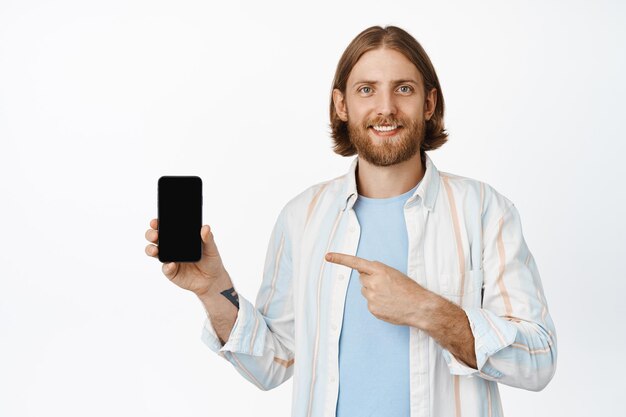 Portrait d'un homme barbu souriant pointant du doigt l'écran du smartphone, l'application d'interface, la publicité de spectacle, l'application en ligne, debout en chemise sur fond blanc